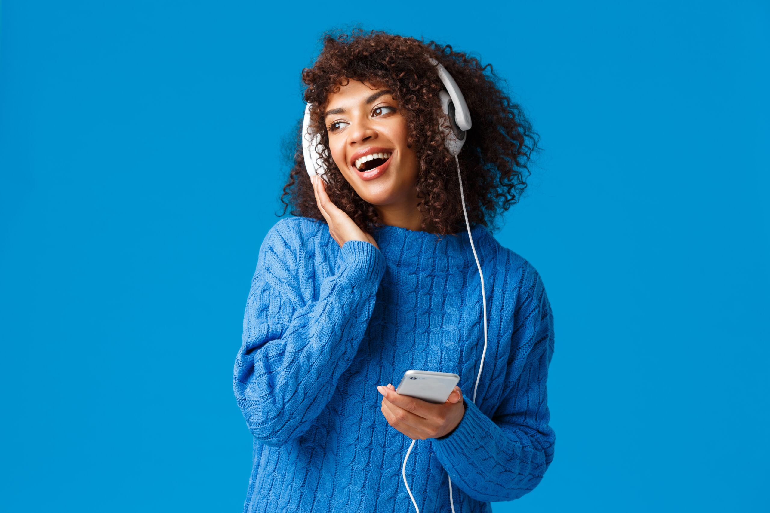 Charismatic modern young attractive african-american girl with afro haircut, listening music in headphones looking out window snowy weather, holding smartphone, smiling and singing